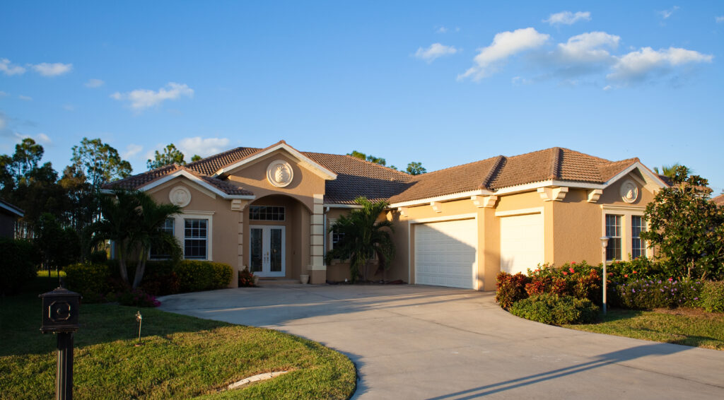 Single-story tan home with a double garage, landscaped lawn, palm trees, and a bright blue sky with scattered clouds.