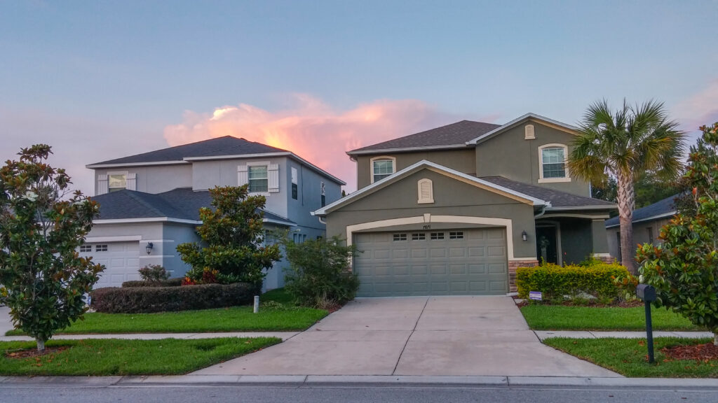 Front view of a two-story suburban home with a driveway, trimmed bushes, palm trees, and a colorful sunset sky overhead
