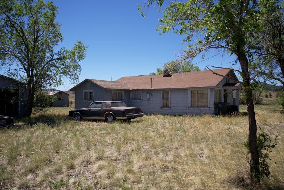 An old, abandoned house with a vintage car in an overgrown yard, situated outdoors.
